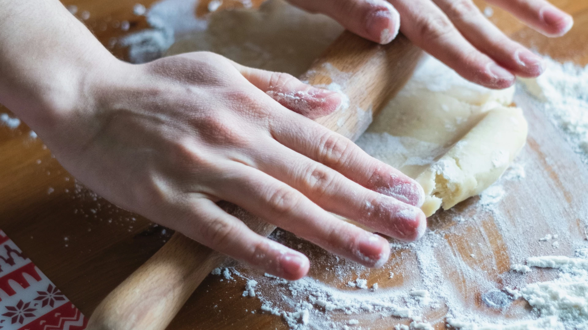 Widok na dłonie ugniatające ciasto wałkiem na drewnianym blacie pełnym mąki.